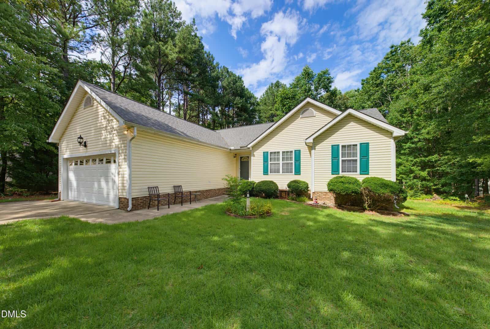 Charming single-story yellow house with green shutters at 95 Wembley Court in Youngsville, featuring an attached two-car garage, a small front porch with chairs, manicured lawn, shrubs, and surrounded by tall trees under a partly cloudy sky.