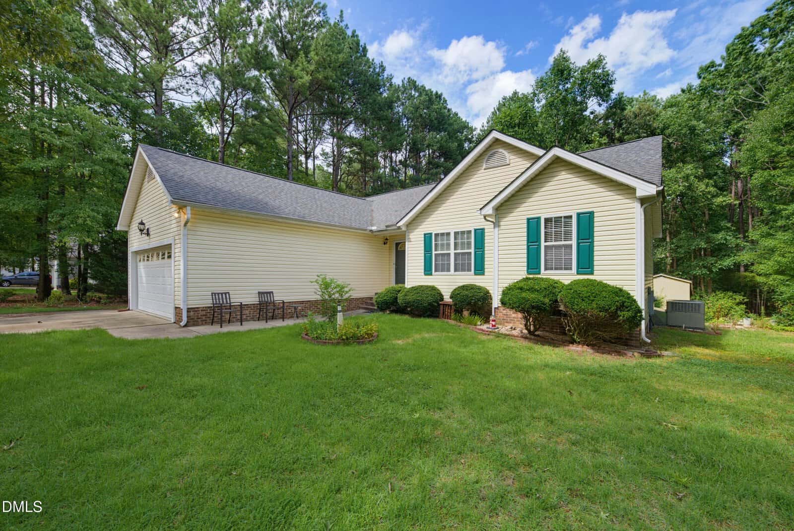 Single-story beige house with green shutters, a two-car garage, and a small front patio with chairs. Surrounded by green grass, bushes, and tall trees under a blue sky with clouds.
