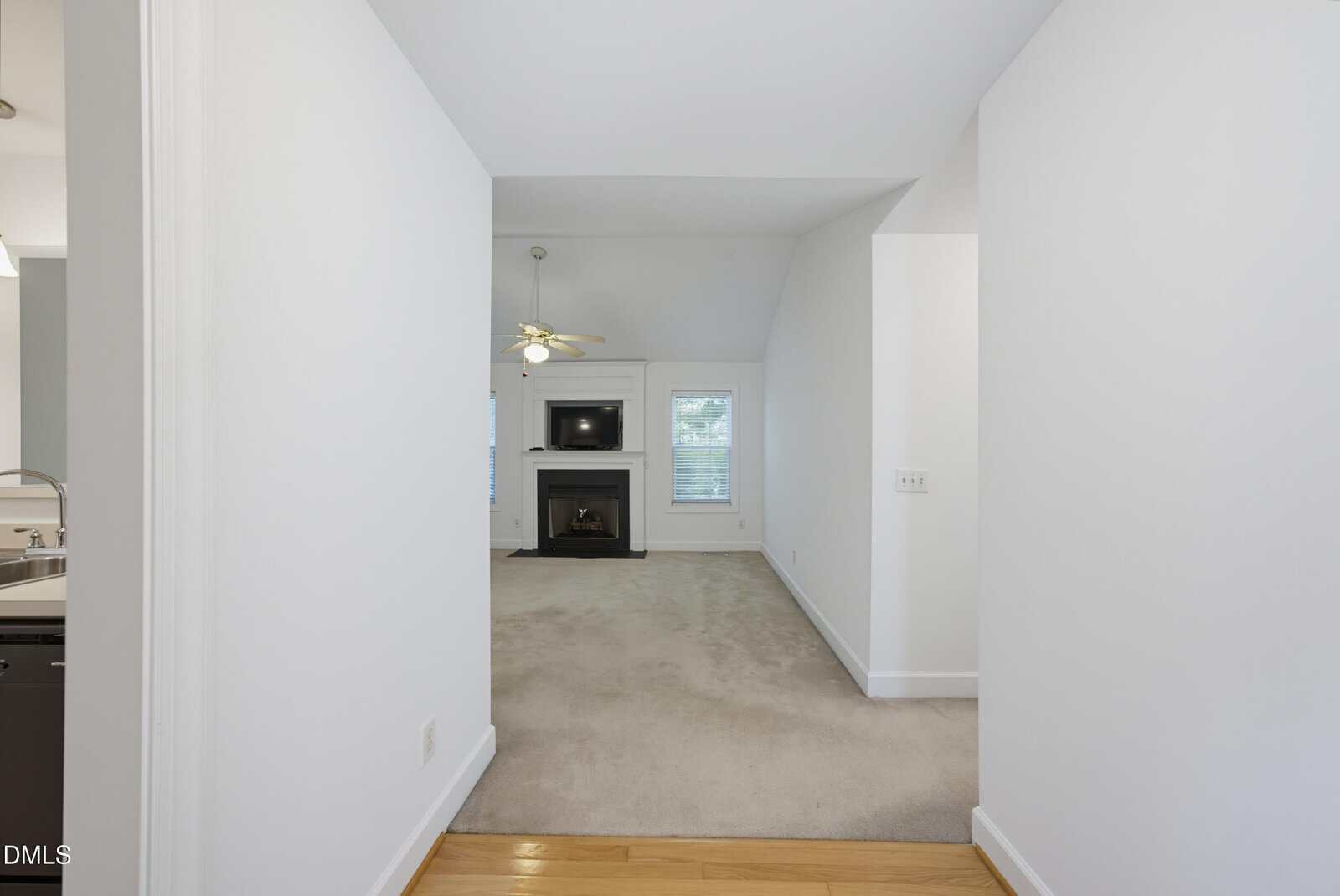 View from a hallway into a living room with beige carpet, a fireplace, a TV above it, windows on either side, and a ceiling fan with lights. A portion of the kitchen counter is visible on the left.