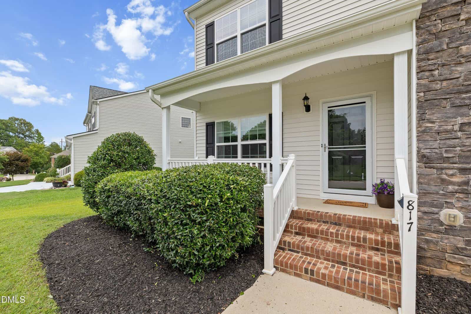 A suburban house with beige siding, brick steps, a white porch, black shutters, and neatly trimmed bushes in front. The house number 817 is visible by the stairs.