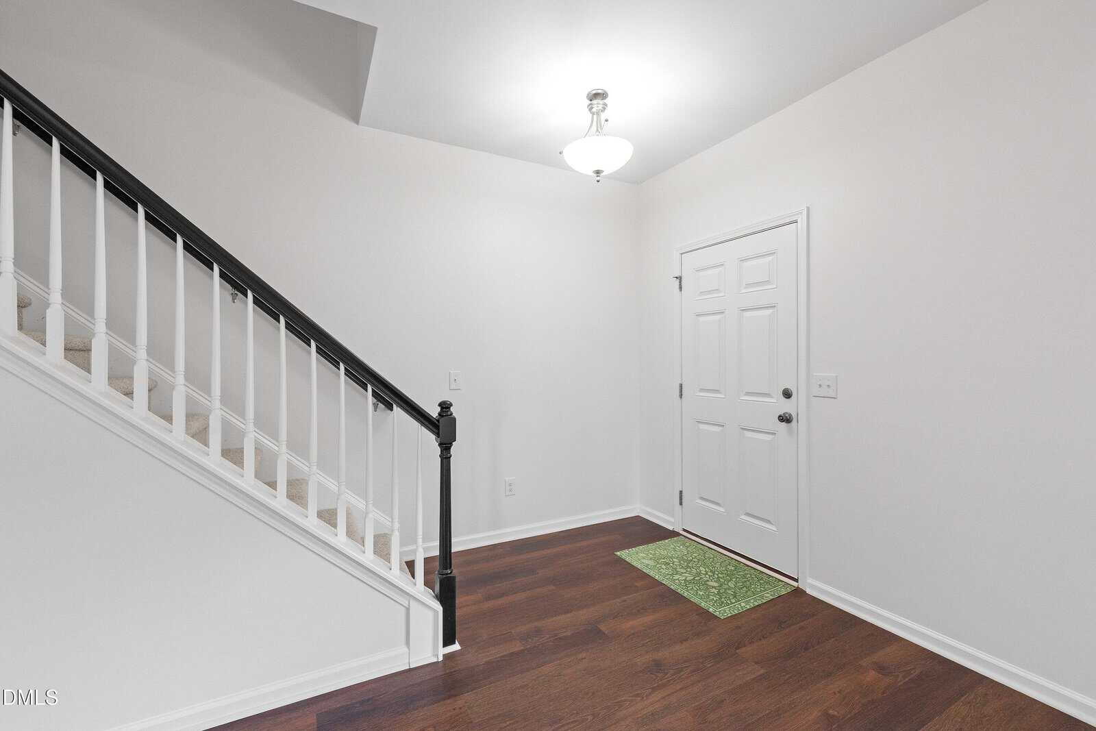 A white front door with a silver handle, a green mat, dark wood flooring, white walls, and a staircase with black railing leading upward. A round ceiling light fixture is above the entryway.
