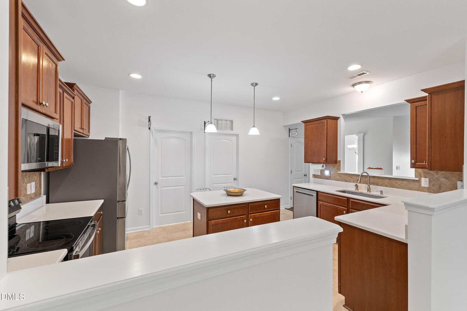 Modern kitchen with wooden cabinets, stainless steel appliances, white countertops, a central island with a bowl, and two pendant lights. Double doors and neutral walls create a clean, bright atmosphere.