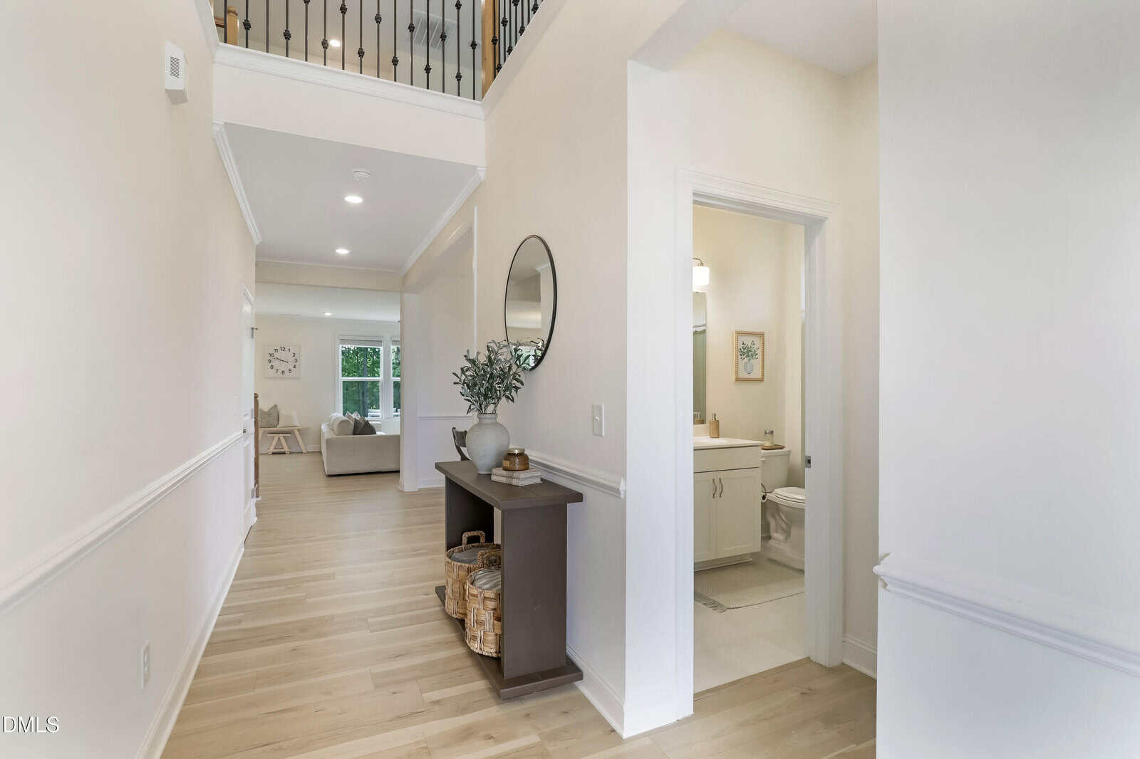 A bright hallway with light wood floors, a console table with decor, a round mirror, and a view into a bathroom and living room. Black railing is visible upstairs.