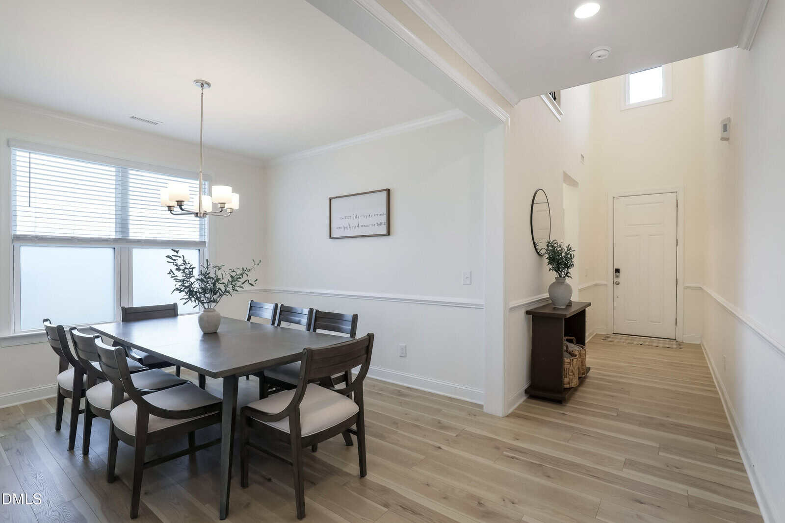 Modern dining room with a dark wood table, six chairs, chandelier, large window, decorative plants, and a hallway leading to a white front door with a console table and round mirror.