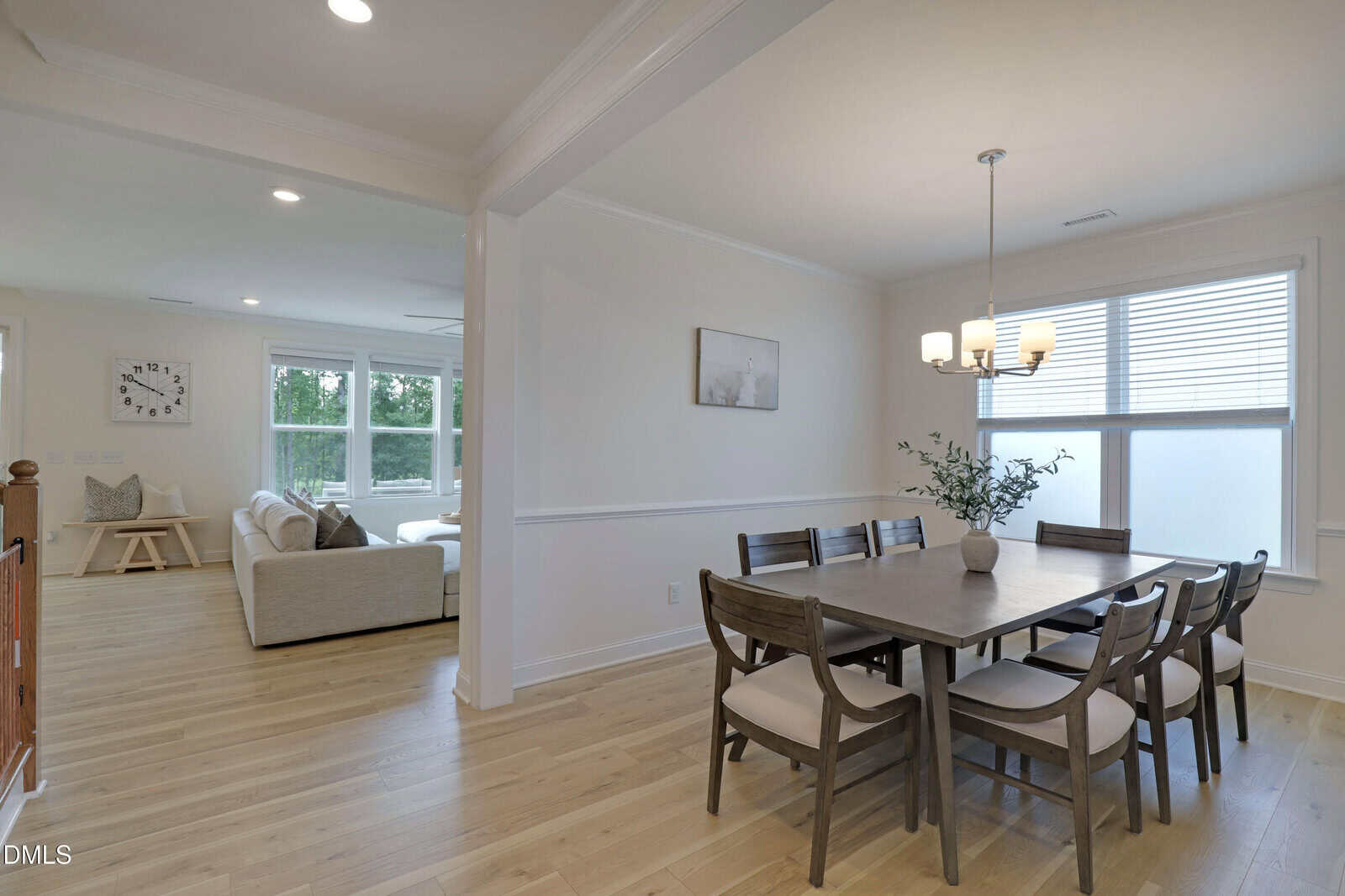 Open dining area with a wooden table and chairs, light fixture above, large window, and adjacent living room with beige sofas and bright natural light.