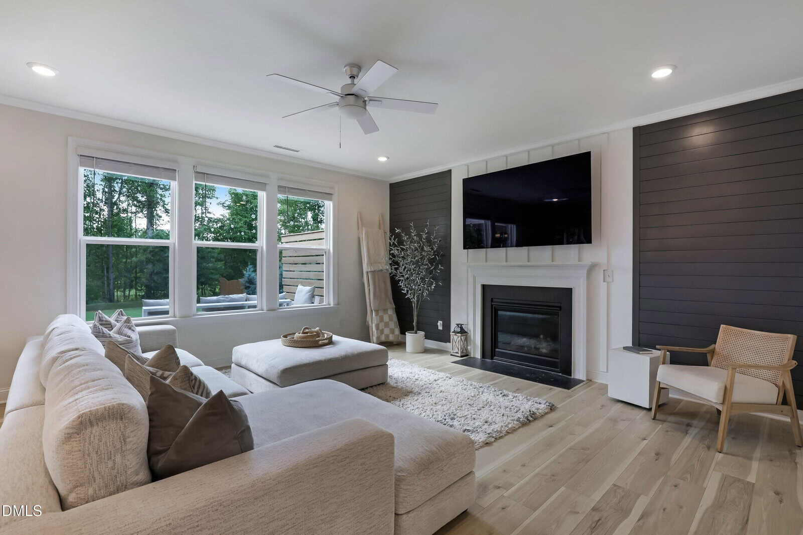 Modern living room with a sectional sofa, ottoman, armchair, fireplace, large wall-mounted TV, shiplap accent walls, wood floors, and big windows showing greenery outside.