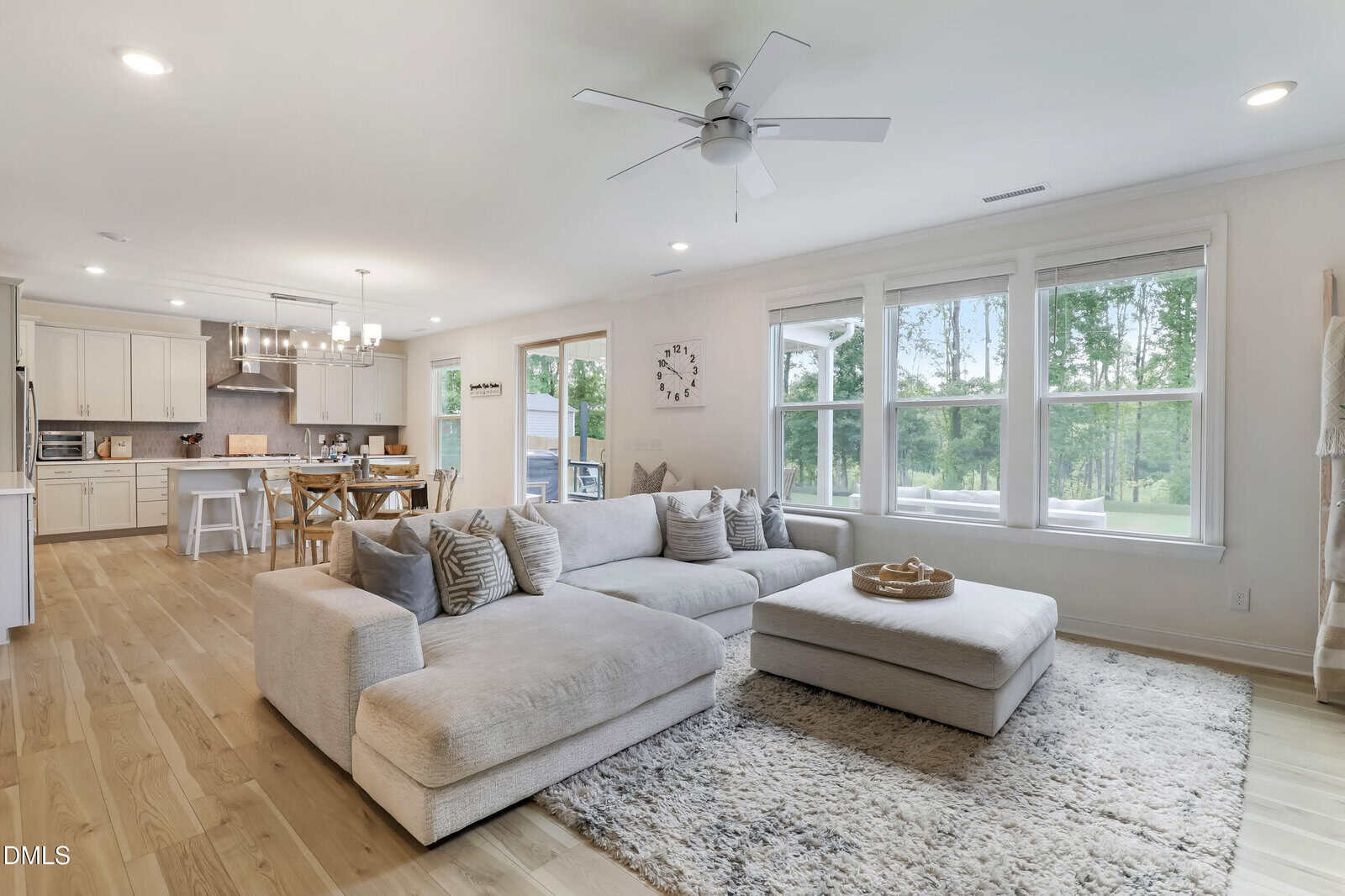 Modern open-concept living room and kitchen with large sectional sofa, ottoman, and rug. Natural light streams in from wide windows, highlighting wooden floors and white cabinetry. A dining table sits near glass doors to the outside.