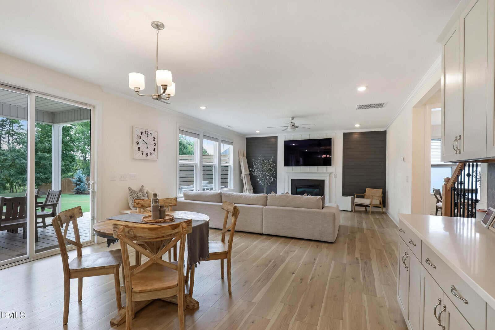 Open-concept living room with light wood floors, a round dining table, beige sofa, wall-mounted TV above a fireplace, large windows, and a door leading to a covered patio with outdoor seating.