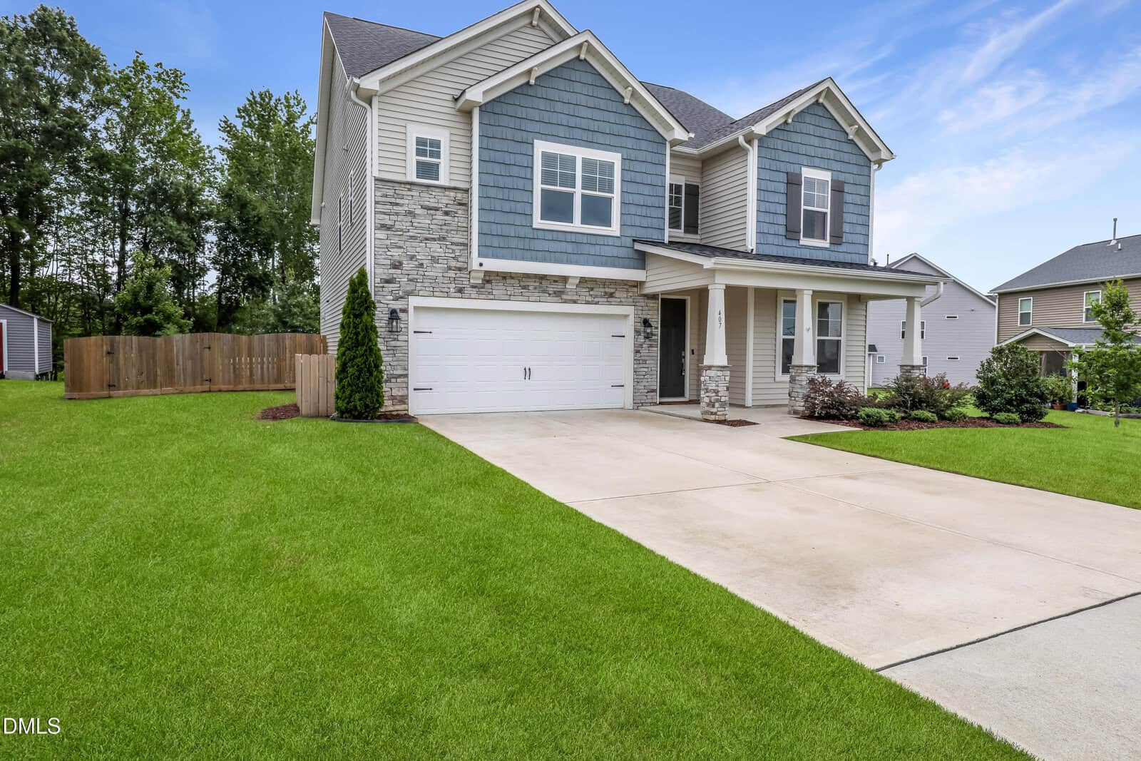 Two-story suburban house with blue and gray siding, white trim, and brick accents. A double garage and a wide driveway lead to a well-maintained lawn and landscaping under a blue sky.