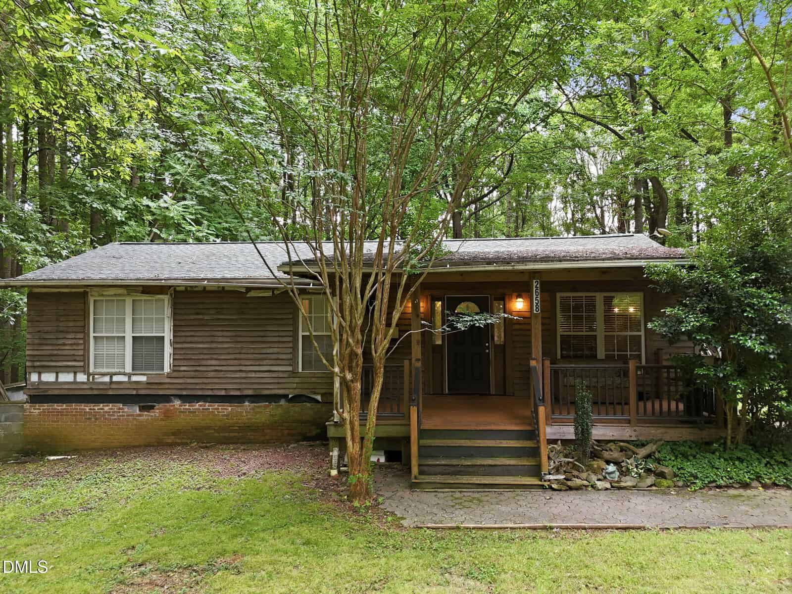 Nestled on a shaded lot at 2658 Goldstone Drive in Oxford, this rustic wooden house features a covered front porch and is surrounded by lush green trees and grass, with a charming stone-lined path leading to the inviting entrance.