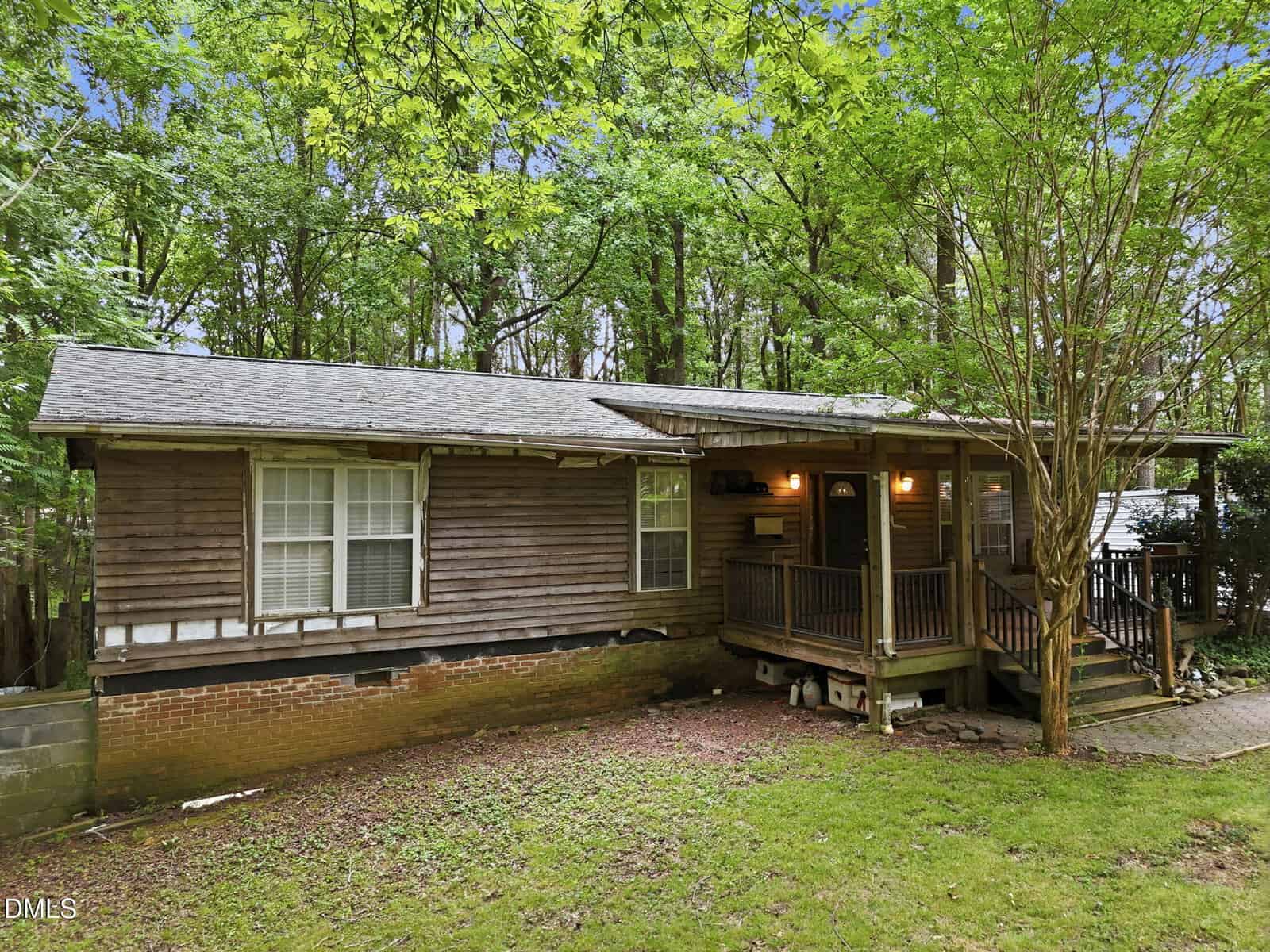A rustic wooden cabin with a covered front porch, surrounded by trees and greenery. The yard is grassy with some bare patches, and there are steps leading up to the porch.