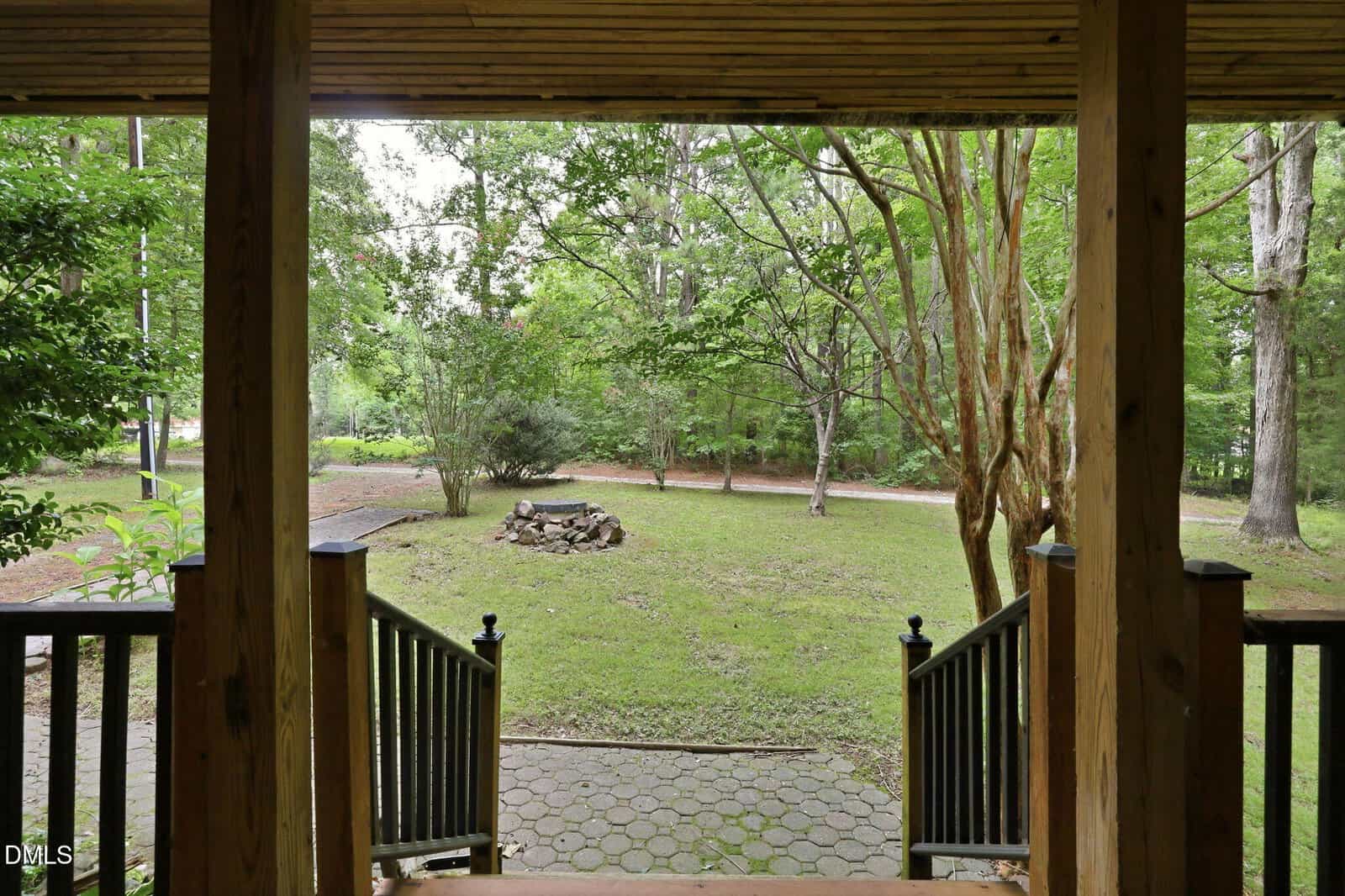 View from a covered wooden porch with steps leading to a green yard, trees, a circular fire pit, and a path. The scene is peaceful and surrounded by nature.