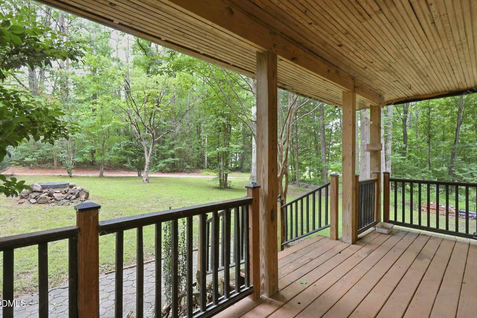 A wooden porch with black railings overlooks a grassy yard bordered by trees. A stone fire pit sits on the lawn, and a paved walkway is visible leading from the porch into the yard.