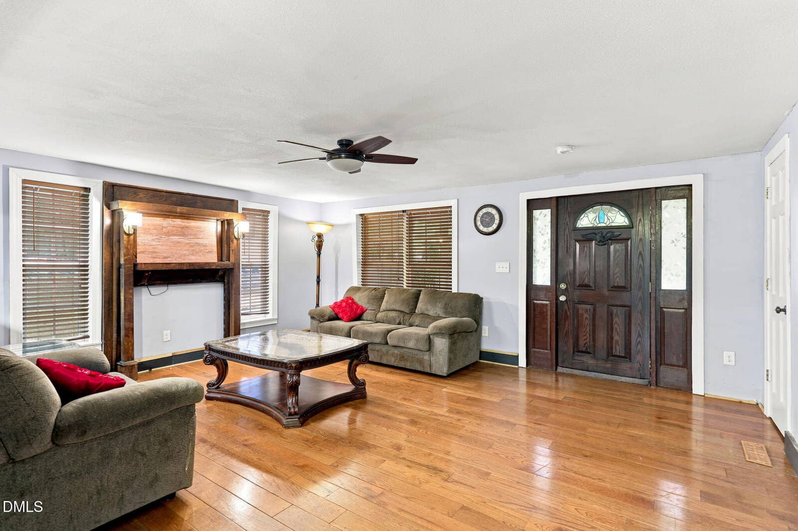 Spacious living room with hardwood floors, two green sofas, a wooden coffee table, fireplace, floor lamp, ceiling fan, and a dark wooden front door with glass panels. Light walls and several windows with blinds.