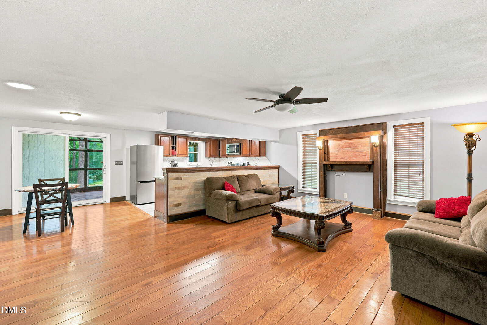 Open living area with wood floors, brown sofas, a glass coffee table, fireplace, and ceiling fan. Kitchen with stainless appliances and dining table visible in the background. Large windows let in natural light.
