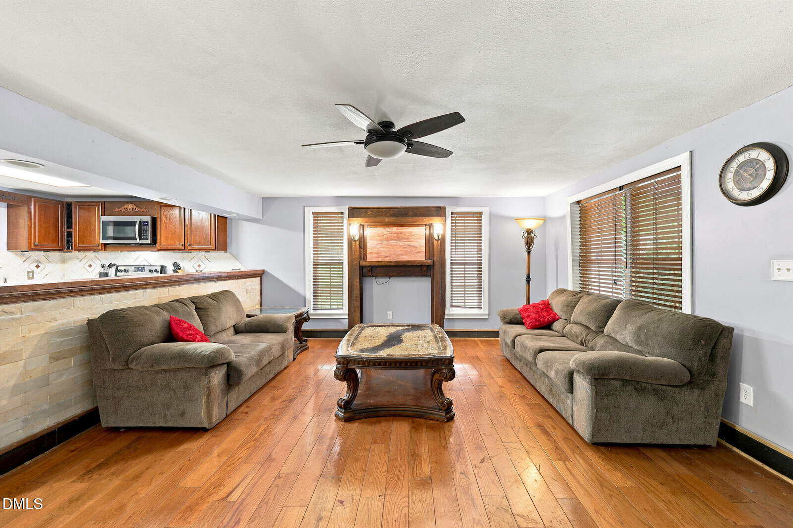 Spacious living room with hardwood floors, two brown sofas, a wooden coffee table, ceiling fan, and large windows. Kitchen with wooden cabinets is visible in the background.