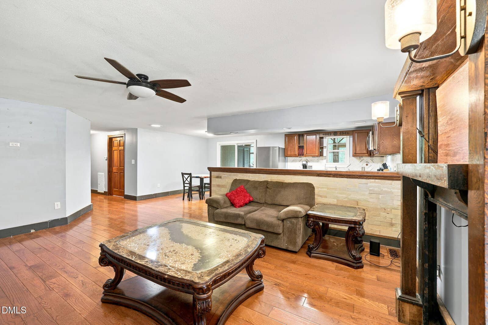Open living room with wood floors, brown sofa, two ornate tables, ceiling fan, and a fireplace on the right. Kitchen with dark cabinets and a breakfast bar is visible in the background. Dining area in the corner.