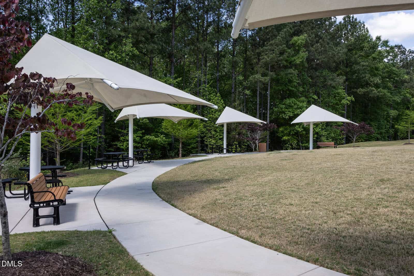 Curved sidewalk with benches and picnic tables under white triangular shade structures in a grassy park area, bordered by trees.