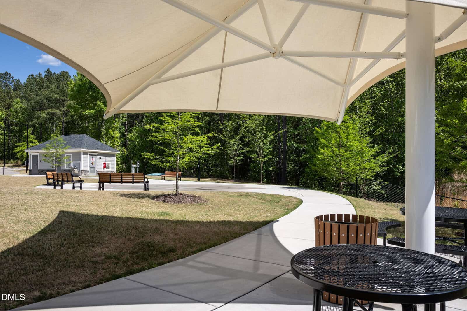 A curved sidewalk passes under a large white shade canopy with black metal tables, near benches, trees, a grassy area, and a small building in the background on a sunny day.