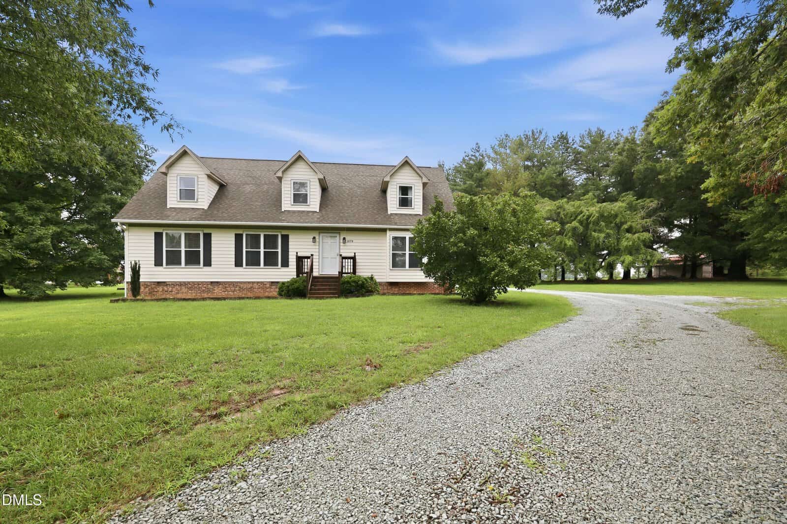 A two-story white house with dormer windows, a brick foundation, and a small front porch, sits on a grassy lawn with a curved gravel driveway and surrounded by trees under a blue sky.