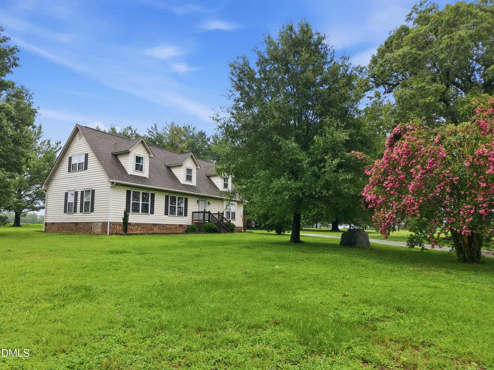 Two-story white house with brown roof, multiple windows, and a brick foundation sits on a green lawn with large trees and pink flowering bushes under a blue sky with scattered clouds.