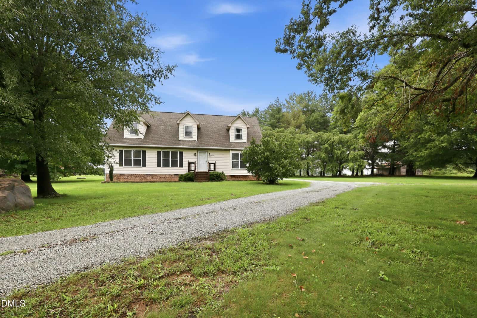 A white two-story house with dormer windows sits on a large green lawn at 2173 Tommie Daniel Road in Oxford. A curved gravel driveway leads to the front of the house, and mature trees surround the property under a blue sky.