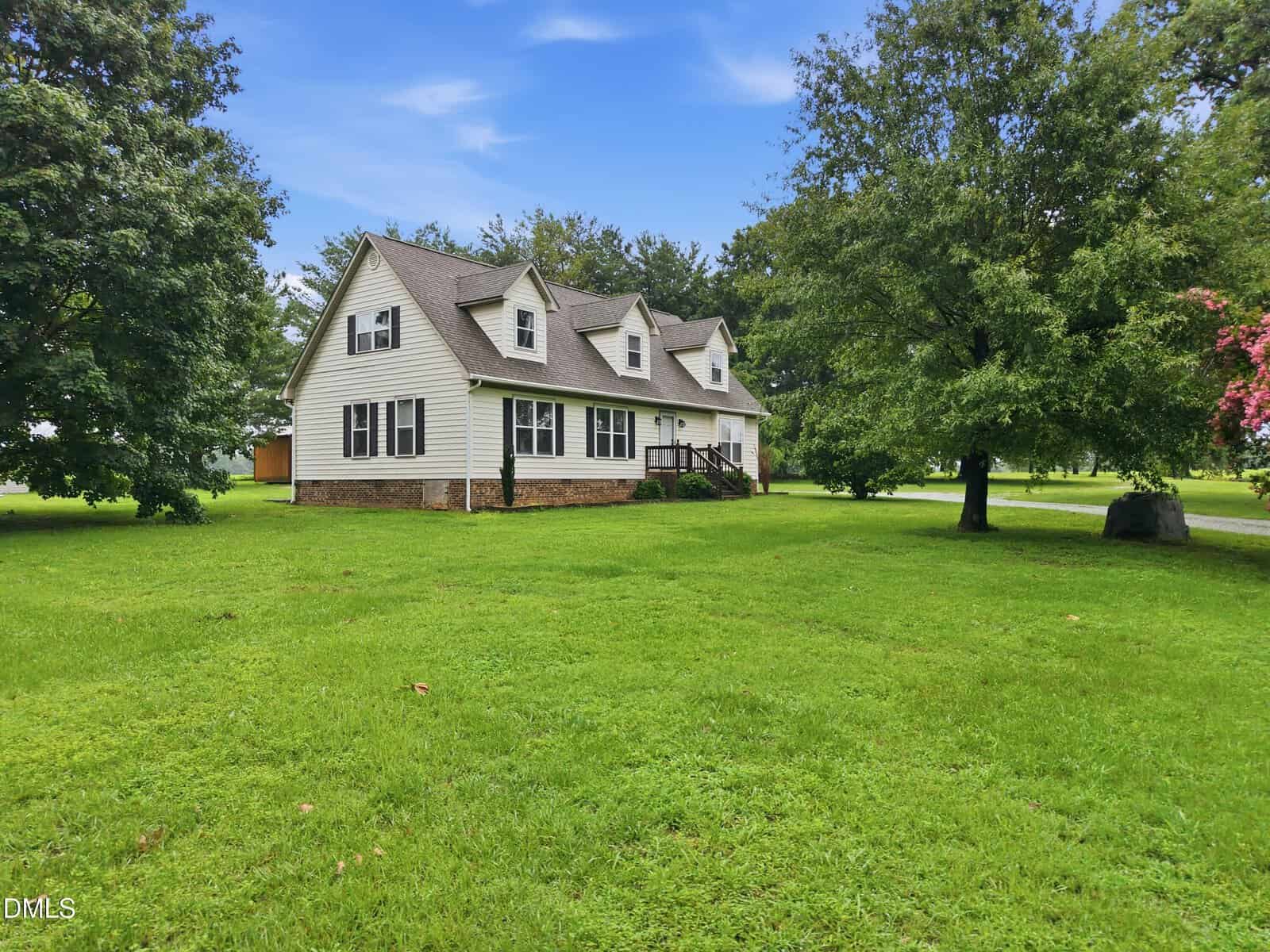 A white two-story house with dormer windows and a brick foundation sits on a large, grassy lawn. Mature trees surround the house, and the sky above is partly cloudy.