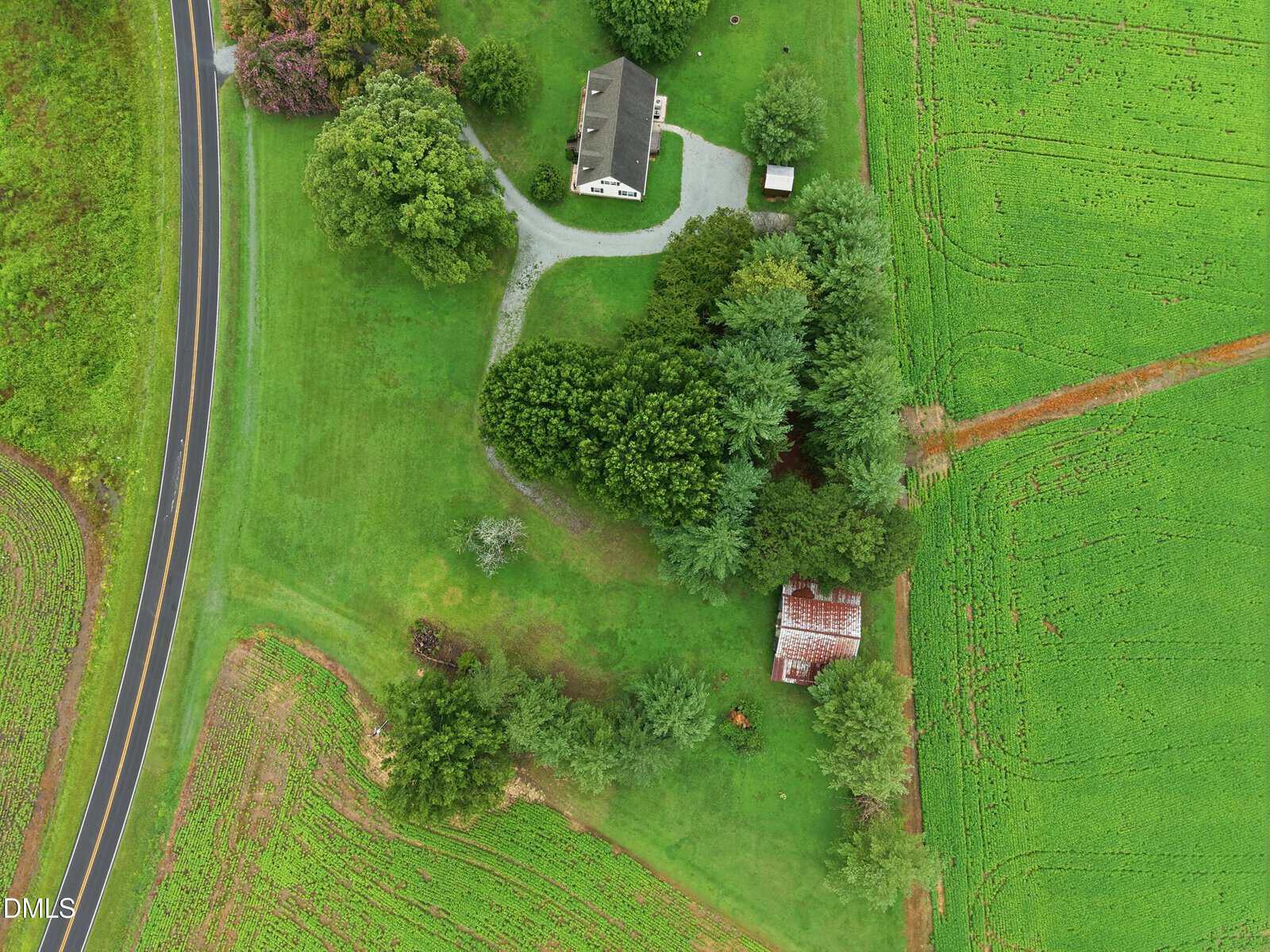 Aerial view of a rural property showing a white house, driveway, garage, trees, and a rusted-roof outbuilding, surrounded by green fields and bordered by a curving road on the left.