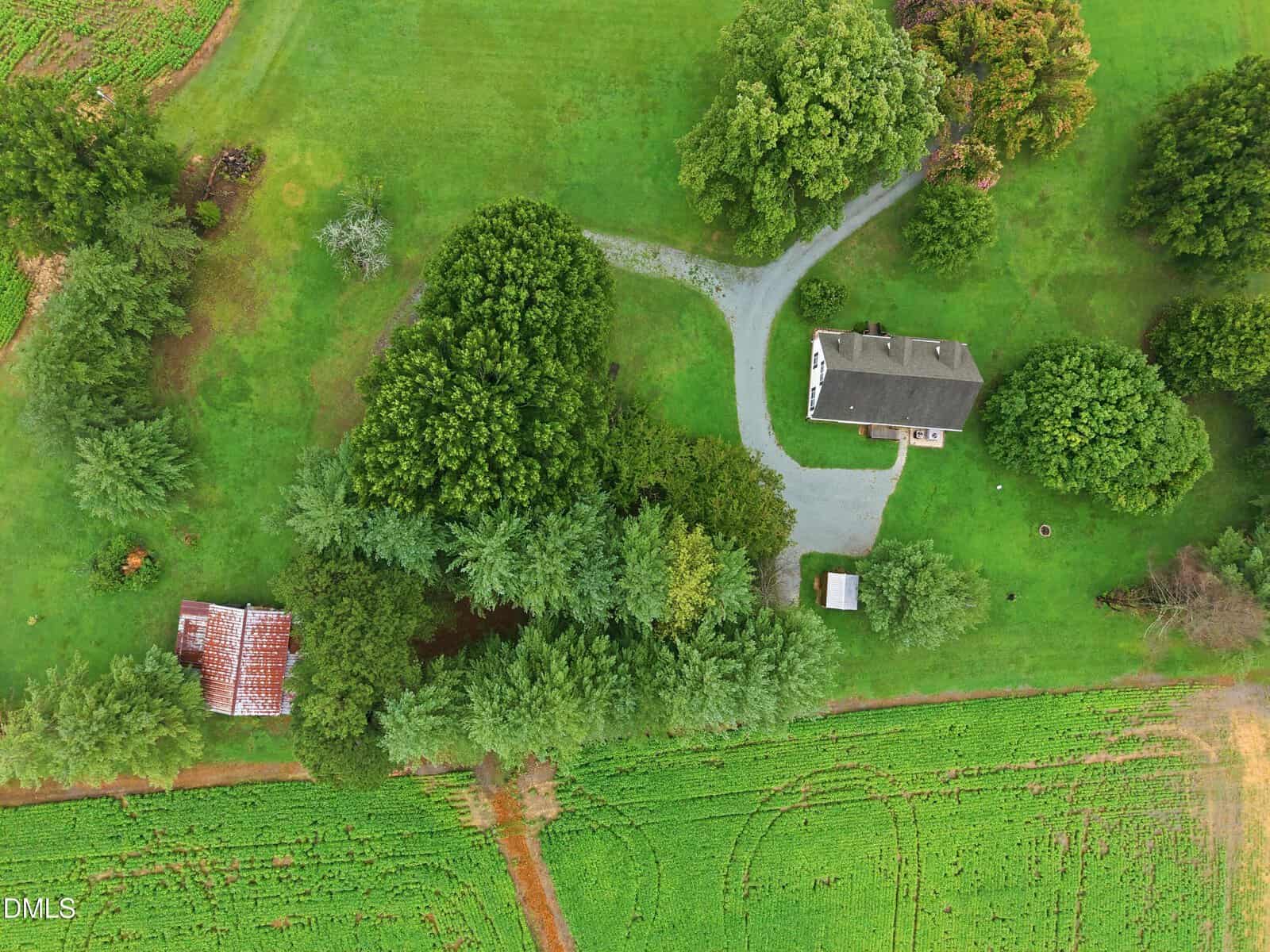 Aerial view of a rural property with a house, a small outbuilding, a gravel driveway, an old rusted-roof structure, and green fields bordered by trees. Paths and farming plots are visible in the surrounding grassy area.