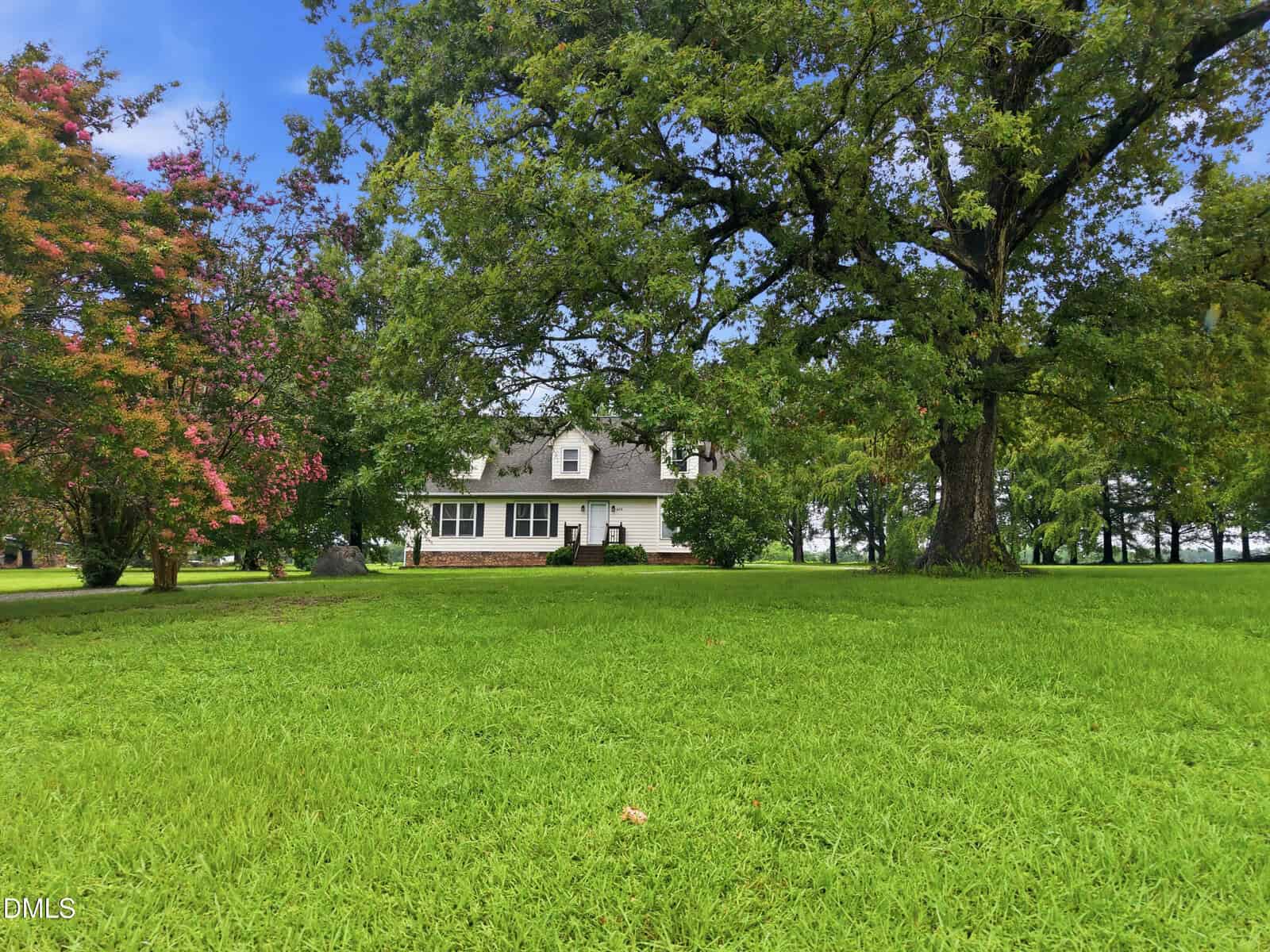 A white two-story house with black shutters is partially visible behind large green trees and a wide, well-manicured lawn under a blue sky with clouds.