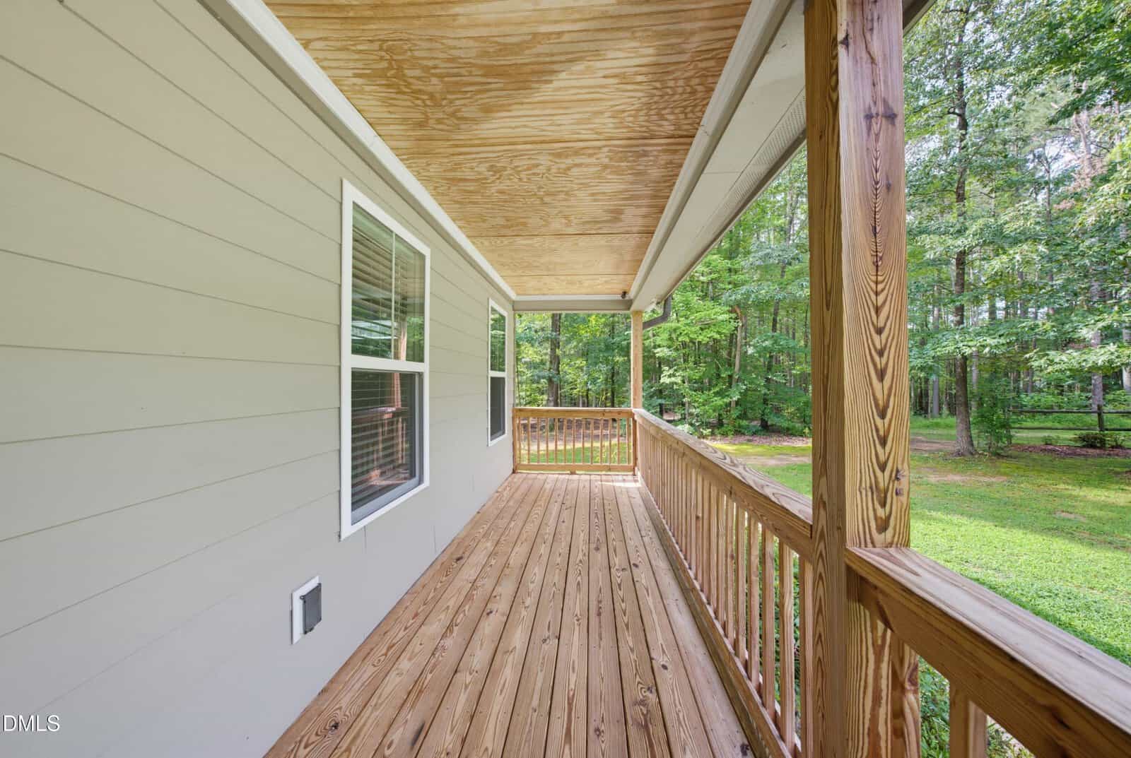 A wooden porch with railings runs alongside a beige house, overlooking a grassy yard and green trees. The porch roof is unfinished wood, and a window is visible on the house wall.