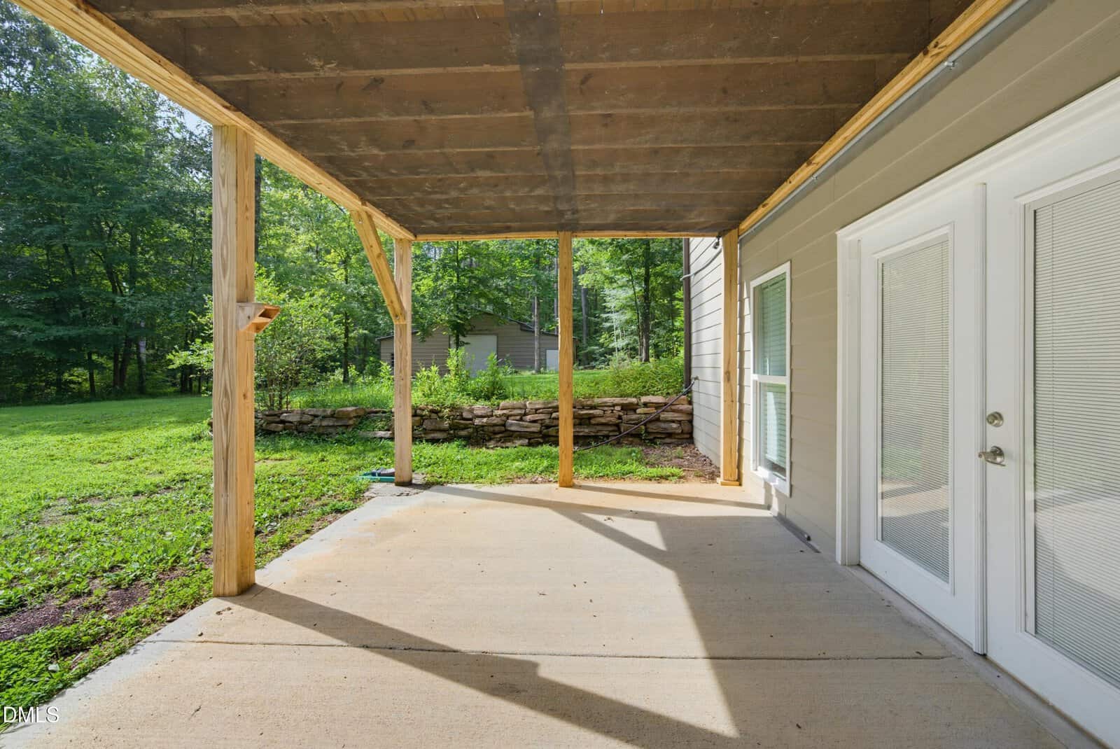 A covered backyard patio with a concrete floor, wooden posts, and double glass doors. Lush green grass, trees, and a stone wall are visible in the background.