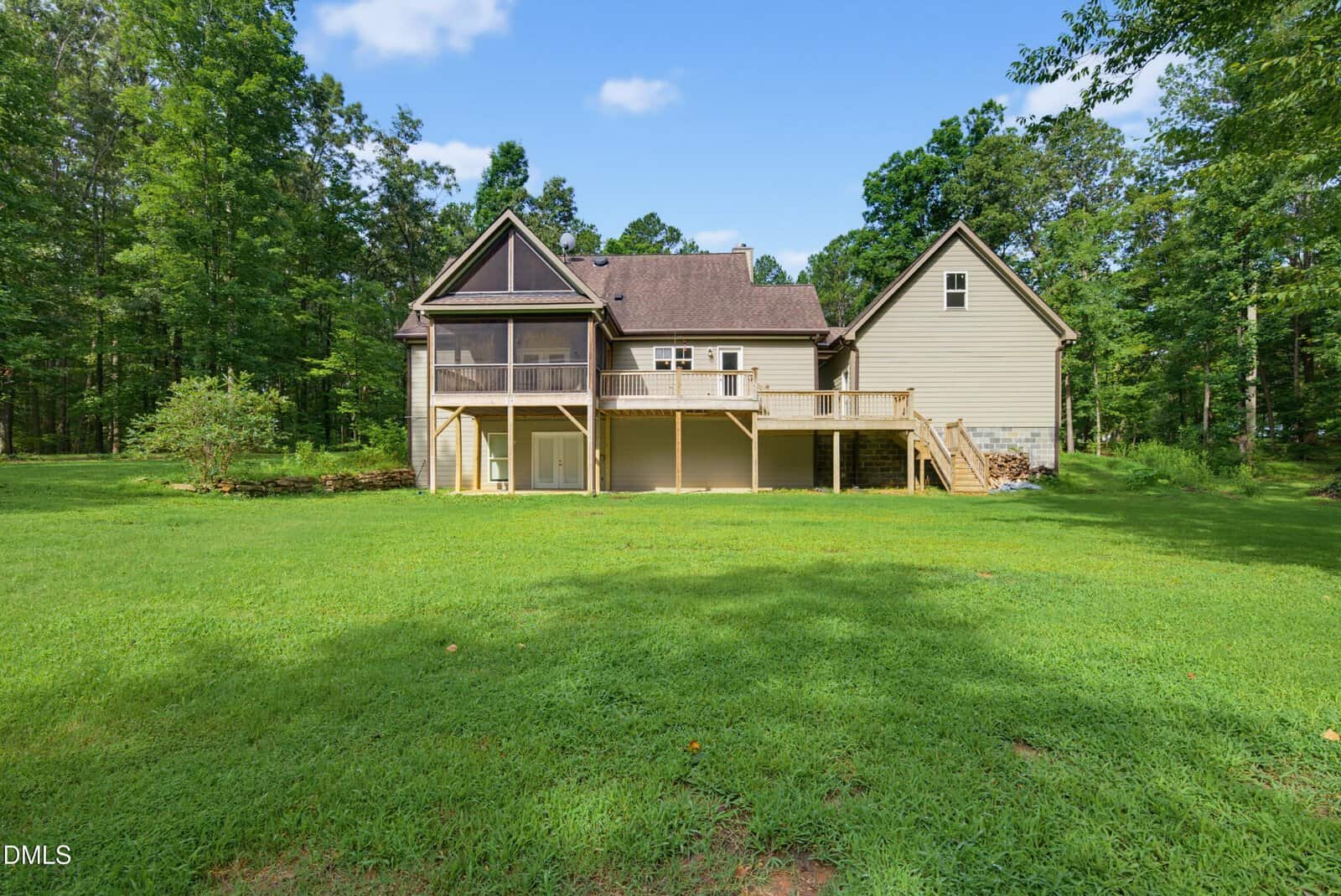 A tan two-story house with a raised wooden deck, screened porch, and stairs leading to a grassy lawn. The home is surrounded by trees and has a large, open backyard under a blue sky with scattered clouds.