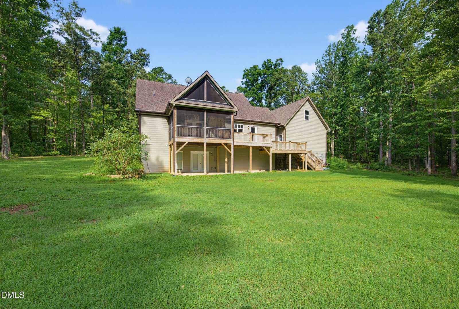 A two-story house with beige siding, a covered porch, and a wooden staircase on a large, grassy lawn surrounded by trees under a clear blue sky.
