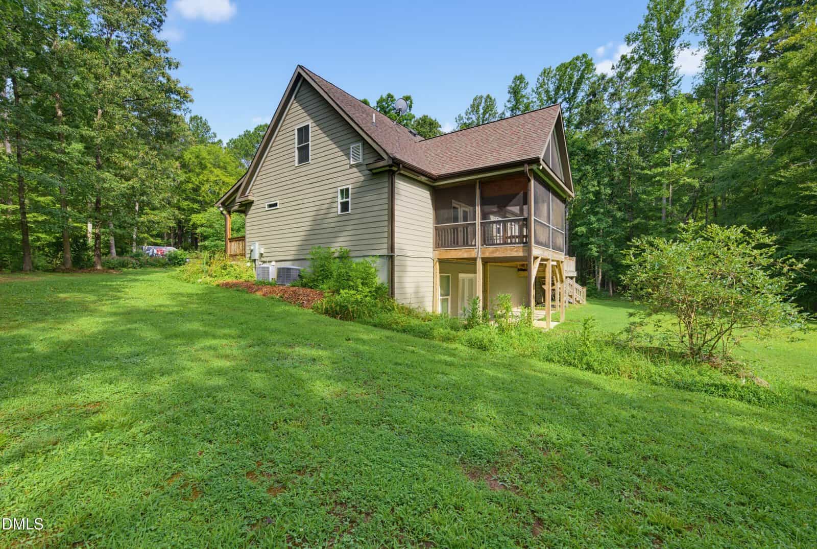 A two-story house with beige siding and a screened porch sits on a grassy lawn, surrounded by dense green trees under a blue sky with scattered clouds.