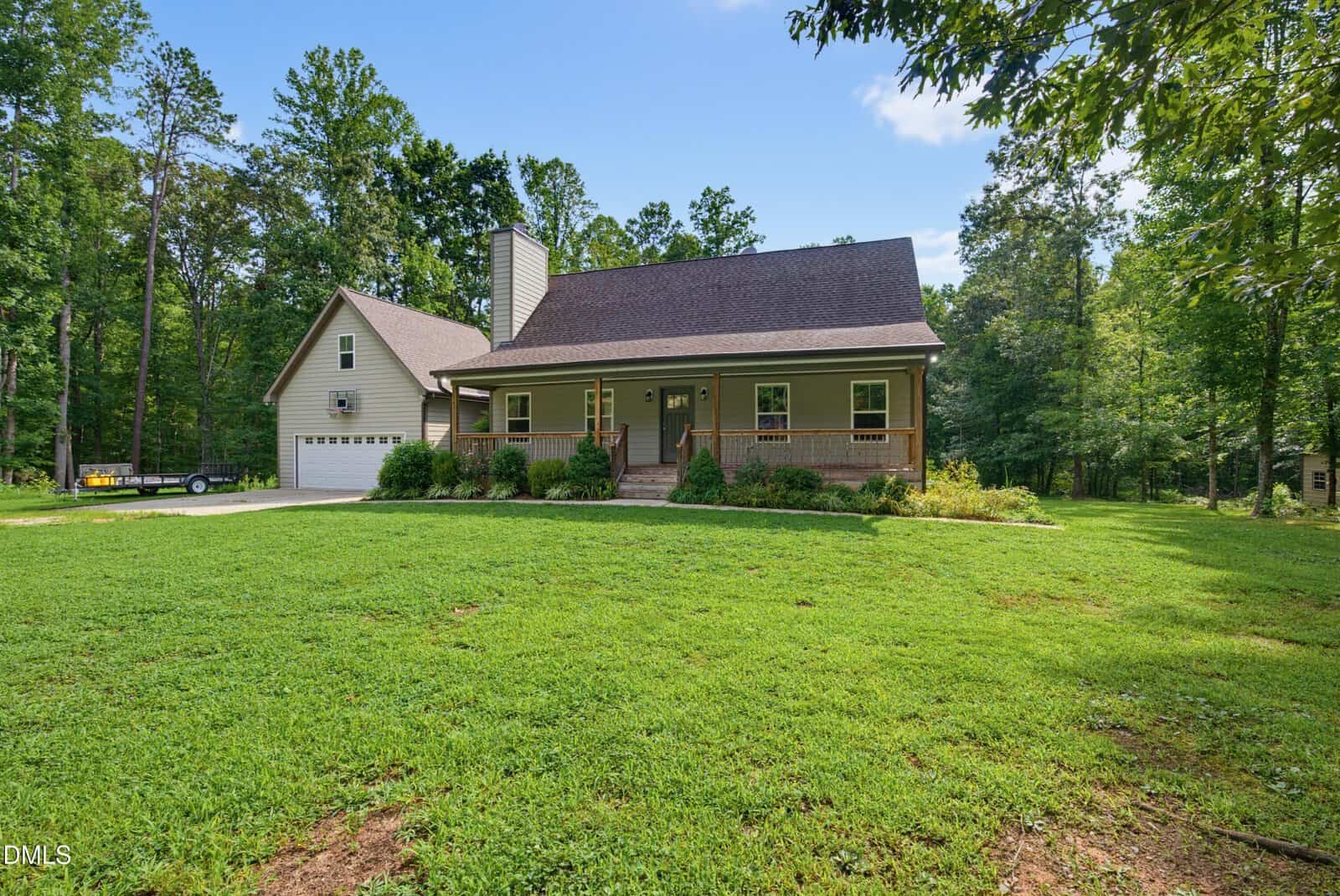 A tan two-story house with a covered porch, attached garage, and a large green lawn surrounded by trees under a blue sky with some clouds.