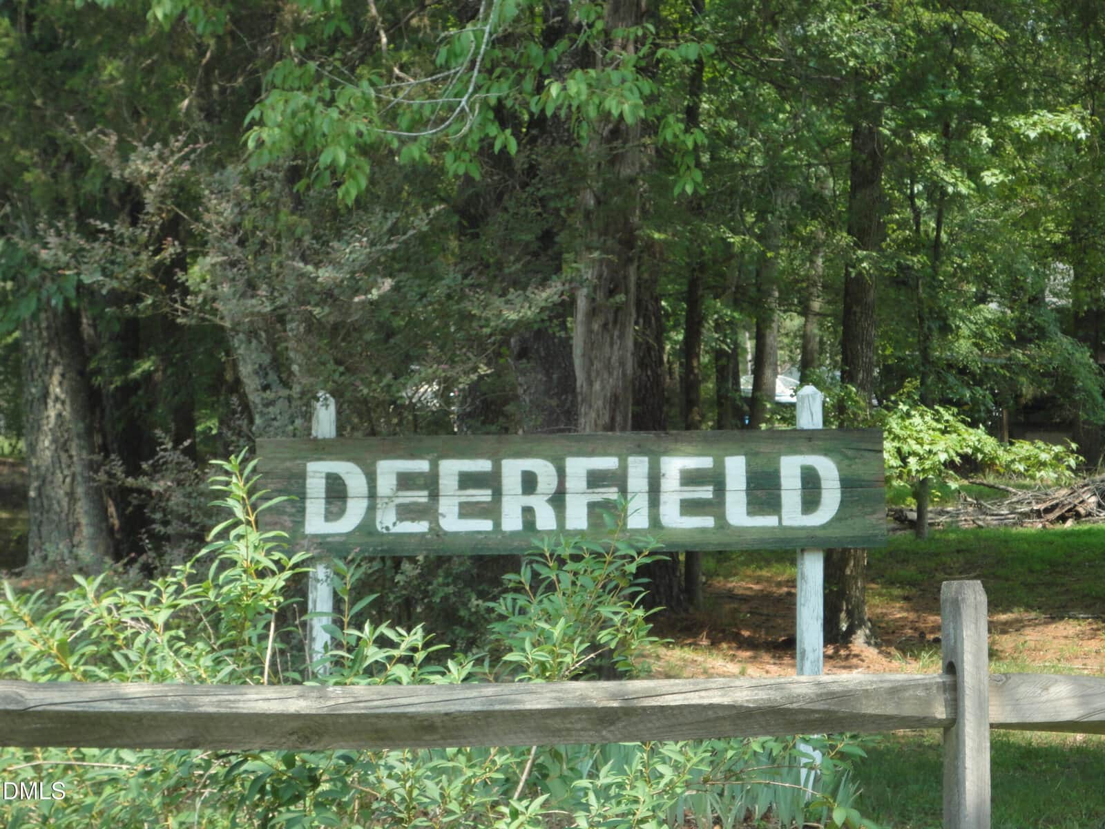 A wooden sign with the word "DEERFIELD" in white letters stands in front of dense trees and greenery at 3588 Deerfield Road, partially obscured by plants, a wooden fence, and clusters of bright green stem.