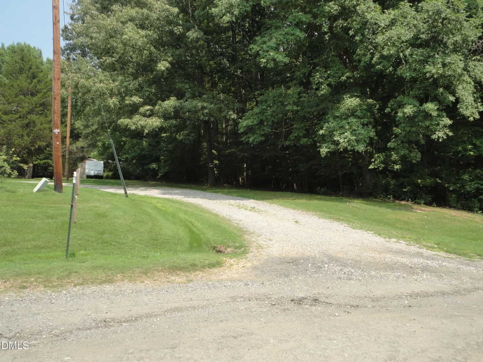 A gravel driveway curves to the right, bordered by grass and trees. Power lines run alongside the drive, and a white vehicle or trailer is partially visible in the background near the tree line.