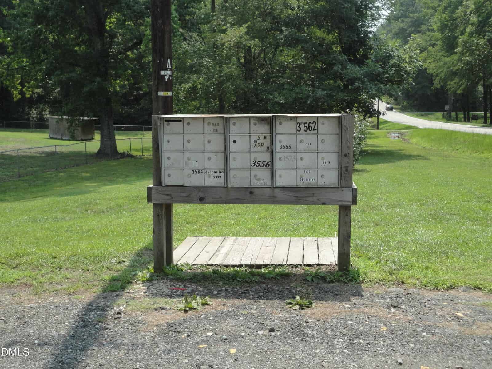 A wooden stand holds multiple metal mailboxes of various sizes by a roadside, with grass, trees, and a street visible in the background. The mailboxes are weathered and numbered.