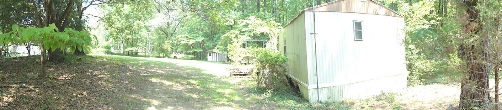 A white mobile home sits on the right side of a shaded, grassy lot surrounded by trees. Sunlight filters through the foliage, and a small shed is visible in the background.