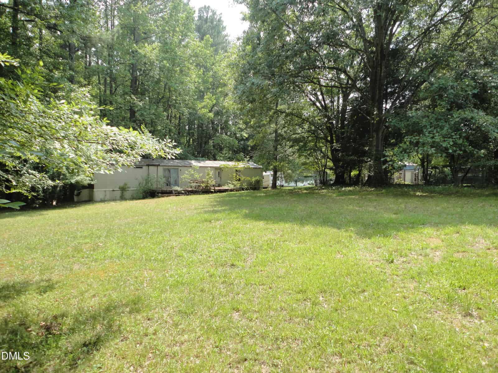 A grassy yard with scattered trees and a white mobile home in the background, surrounded by lush greenery under a sunny sky.