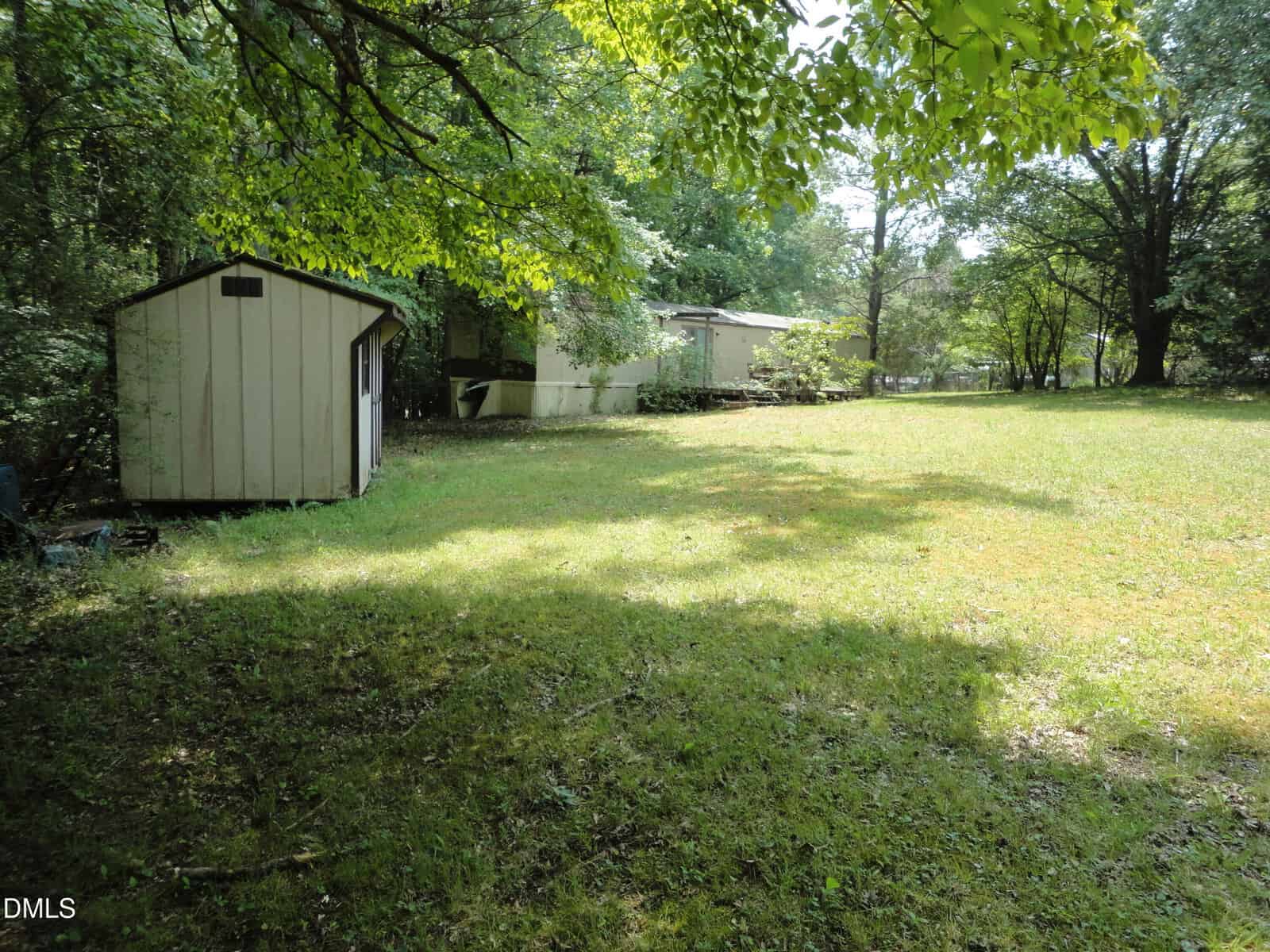 A small shed sits on a grassy yard surrounded by trees, with more trees and a larger building visible in the background under bright sunlight.