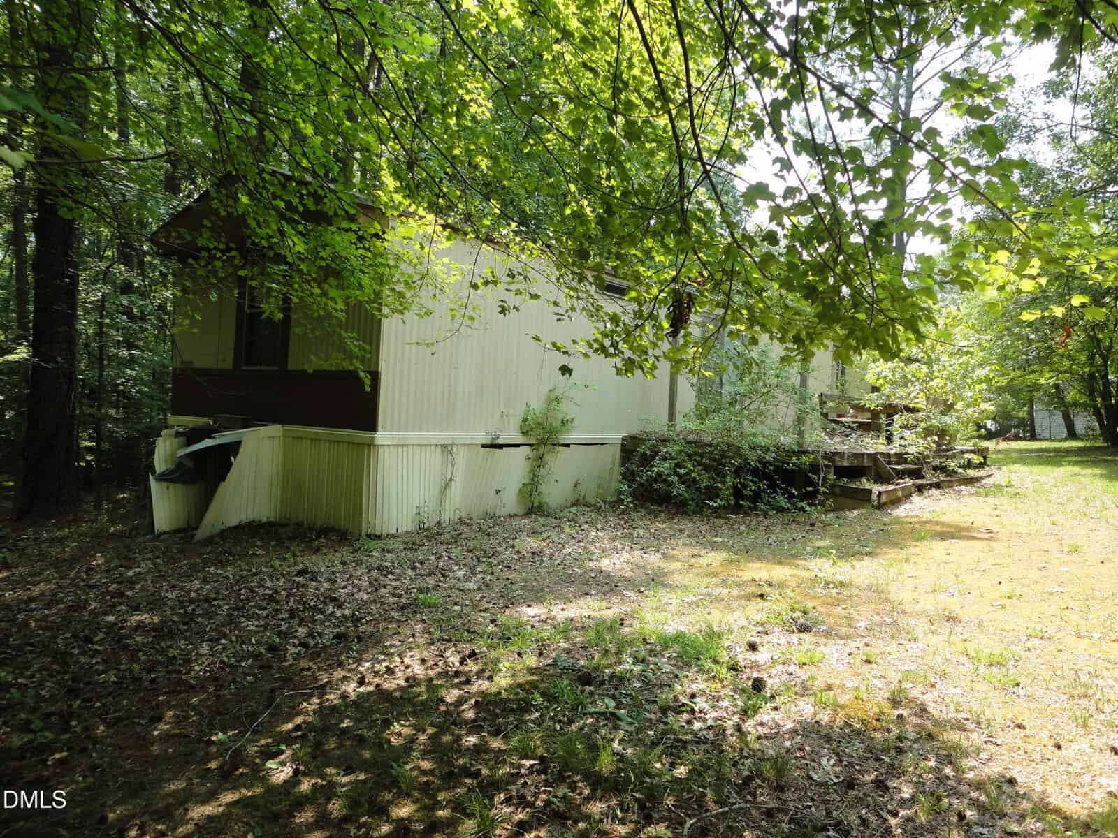 A beige mobile home with a small porch sits in a wooded area, surrounded by green trees and patches of sunlight on the ground.