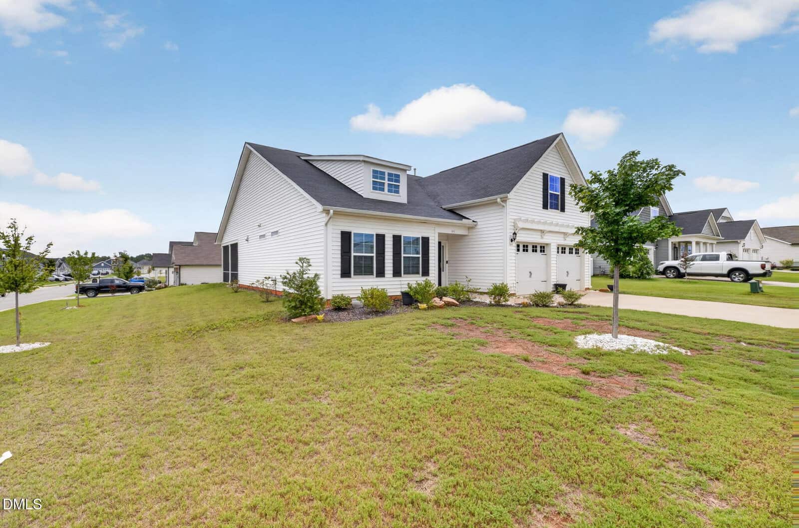 A white two-story suburban house with a dark roof, attached garage, and a small tree in the front yard. The house is surrounded by grass and neighboring homes are visible in the background under a partly cloudy sky.