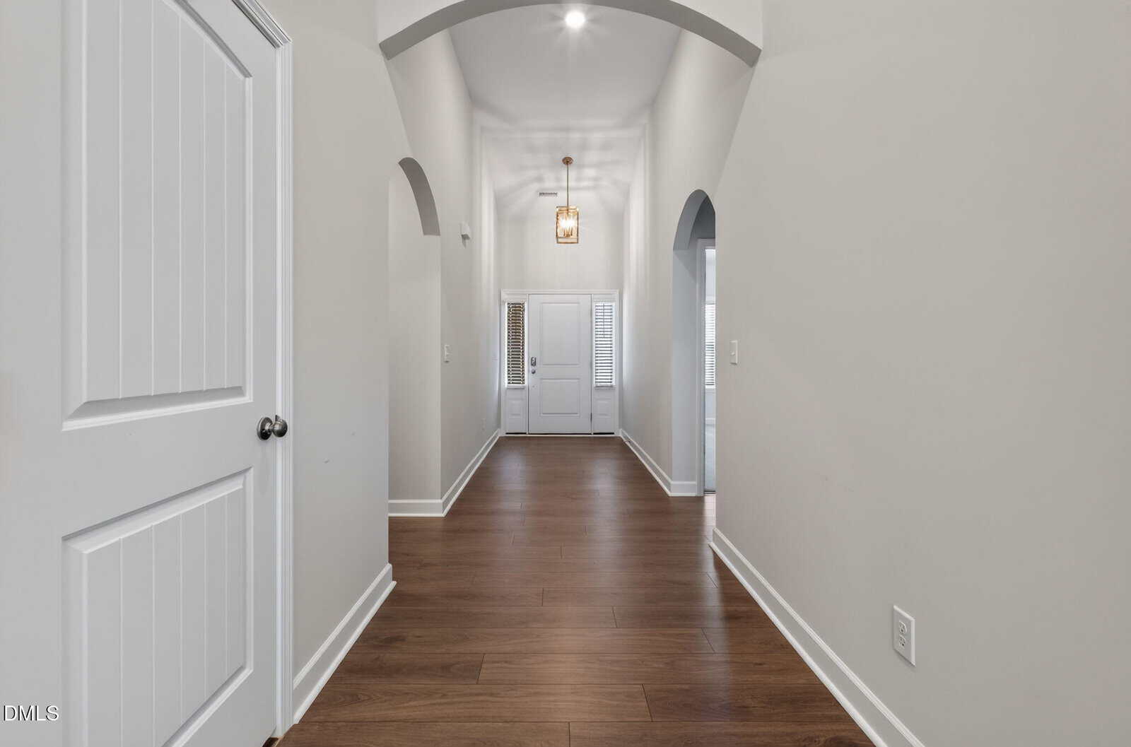 A hallway with light gray walls, wood flooring, an arched ceiling, a white door on the left, and double front doors with glass panels at the end. A modern pendant light hangs from the ceiling.