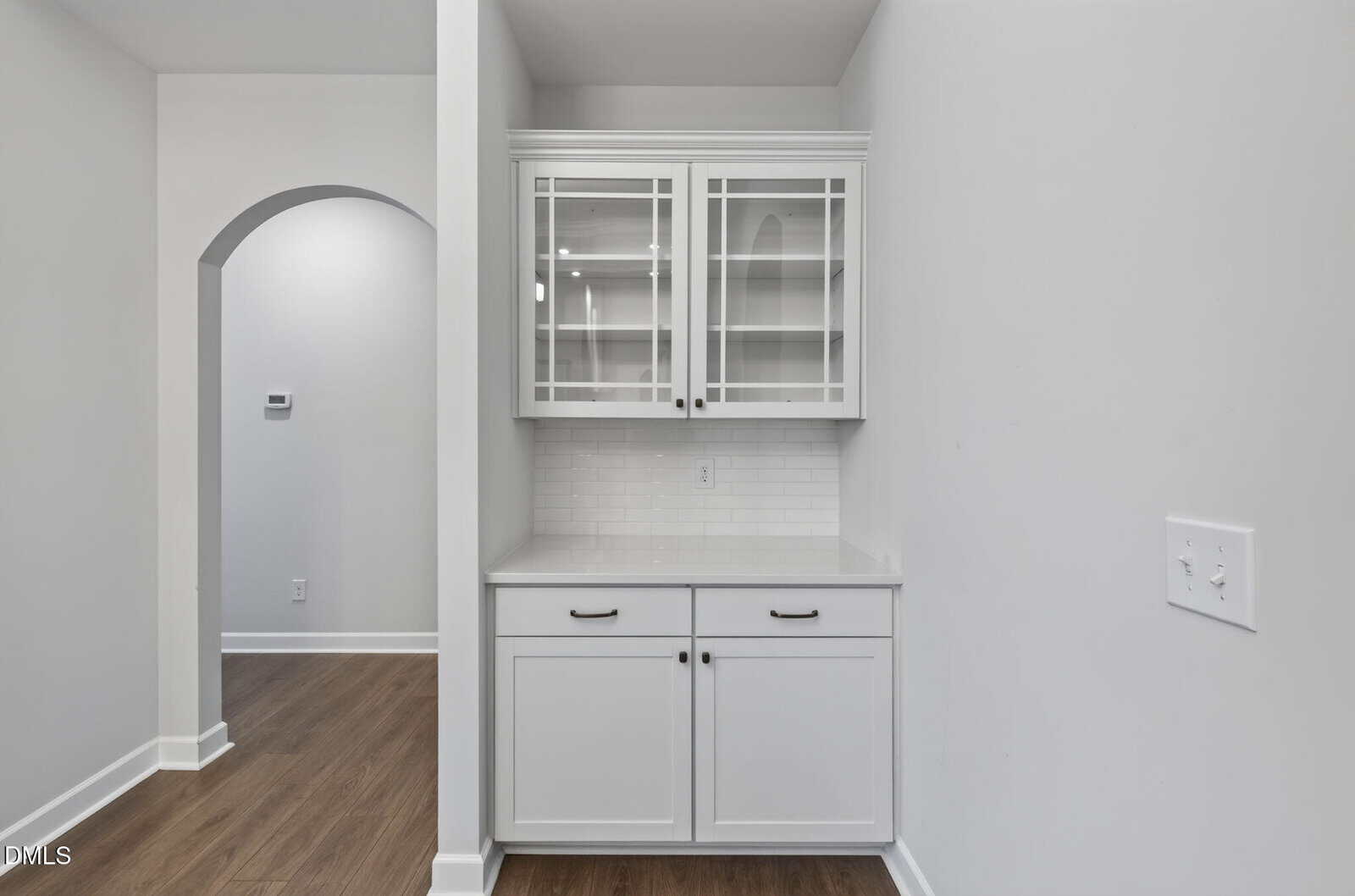 White built-in cabinet with glass upper doors and solid lower doors, featuring black handles. The cabinet is set against a light gray wall near an arched doorway and a light switch, with hardwood flooring.