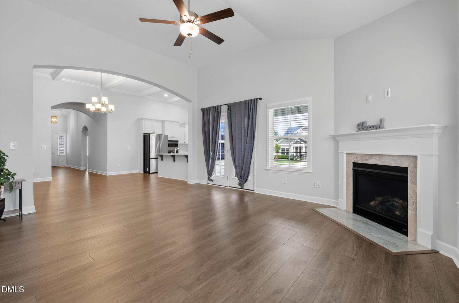 Spacious living room with light wood flooring, a ceiling fan, white walls, a fireplace with a mantel, large window with curtains, and a view into the kitchen and hallway.