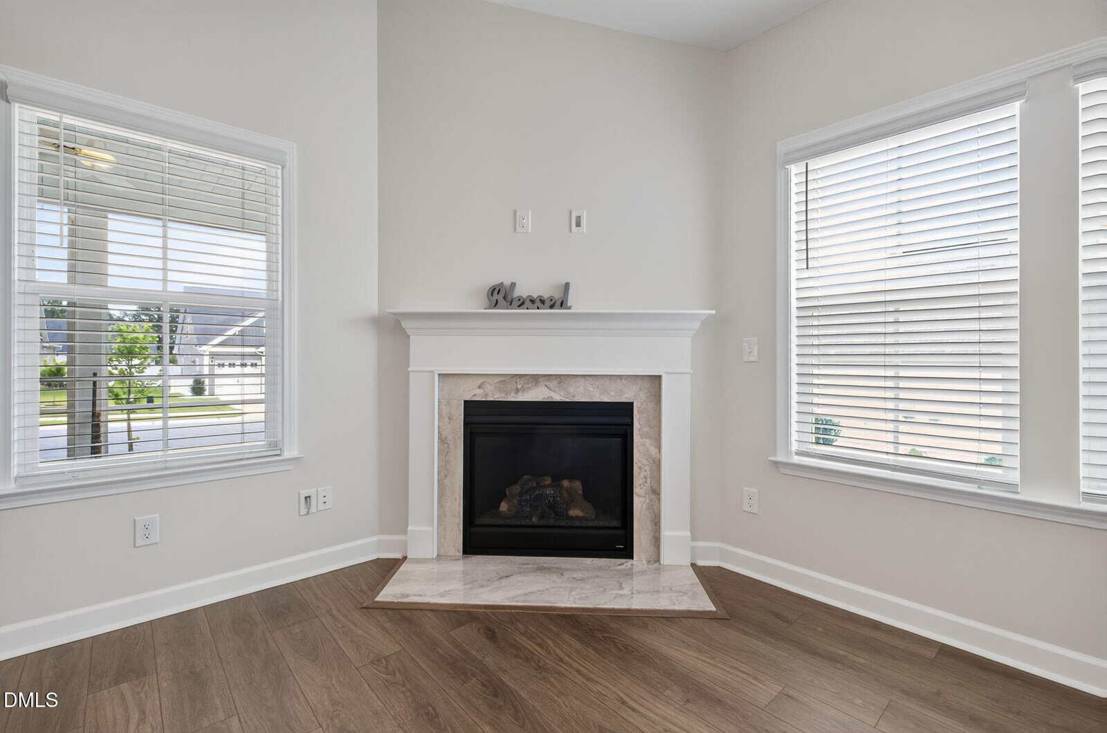 A bright corner room features a fireplace with a white mantel, flanked by two large windows with blinds. The floor is wood, and a small “blessed” sign sits on the mantel.