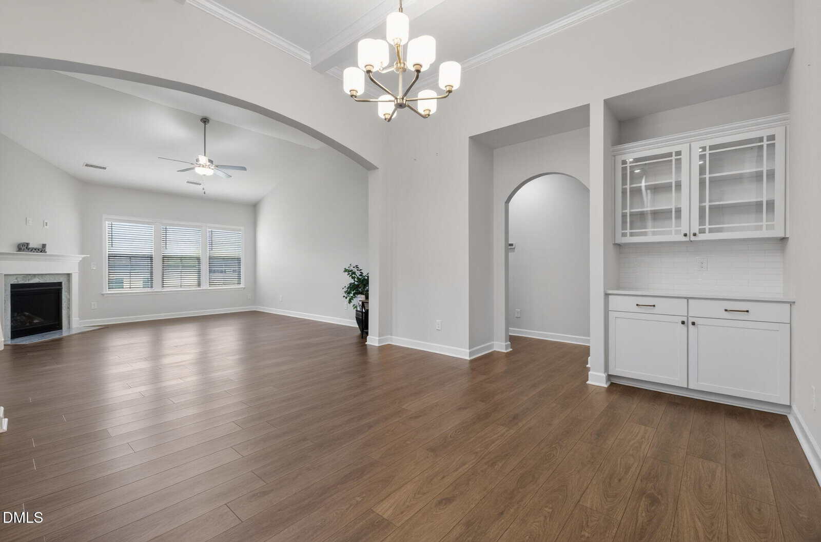 Open living and dining area with wood floors, white walls, a built-in cabinet, chandeliers, large windows, a ceiling fan, and a fireplace in the corner. Arched doorways add architectural interest.