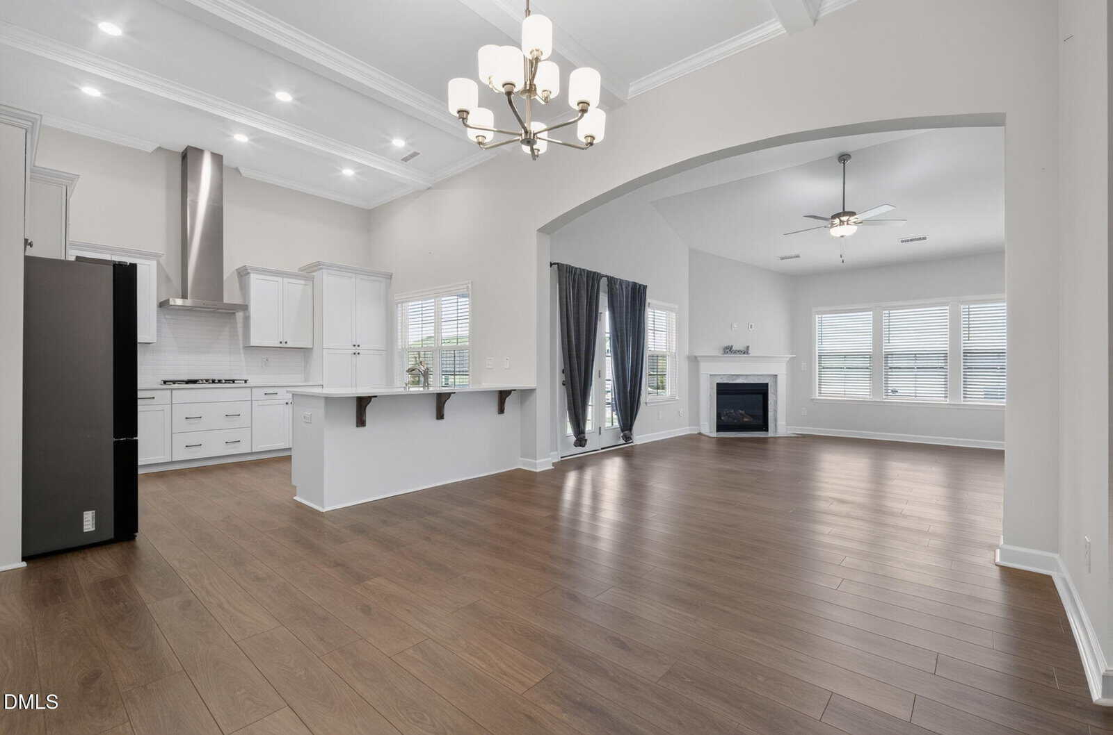 Open-concept kitchen and living room with wood floors, white cabinets, a large island, modern lighting, stainless steel appliances, a fireplace, and windows with gray curtains letting in natural light.